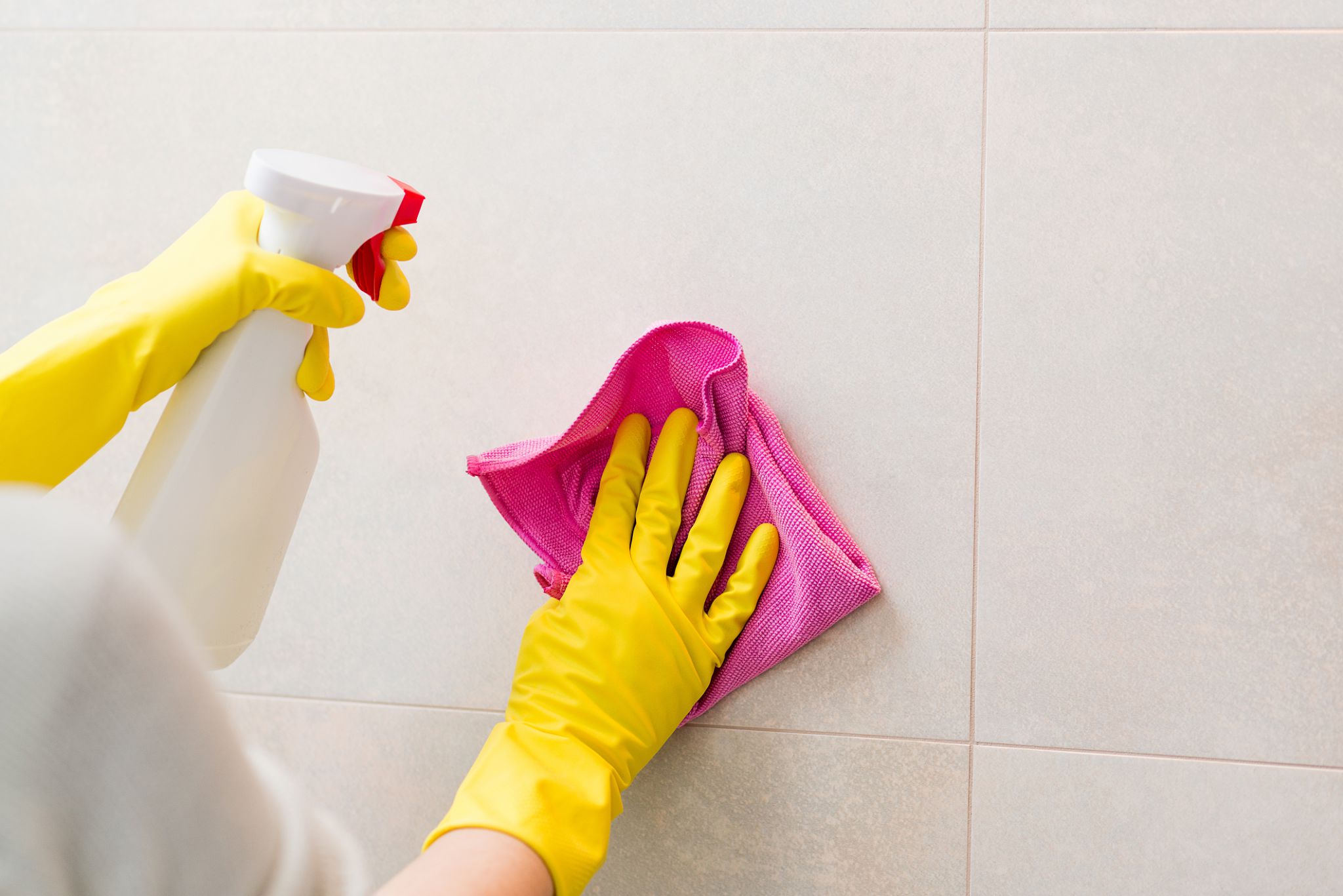 Hands with yellow rubber gloves holding detergent spray bottle and cleaning tiles in bathroom with pink cloth. Spring cleaning